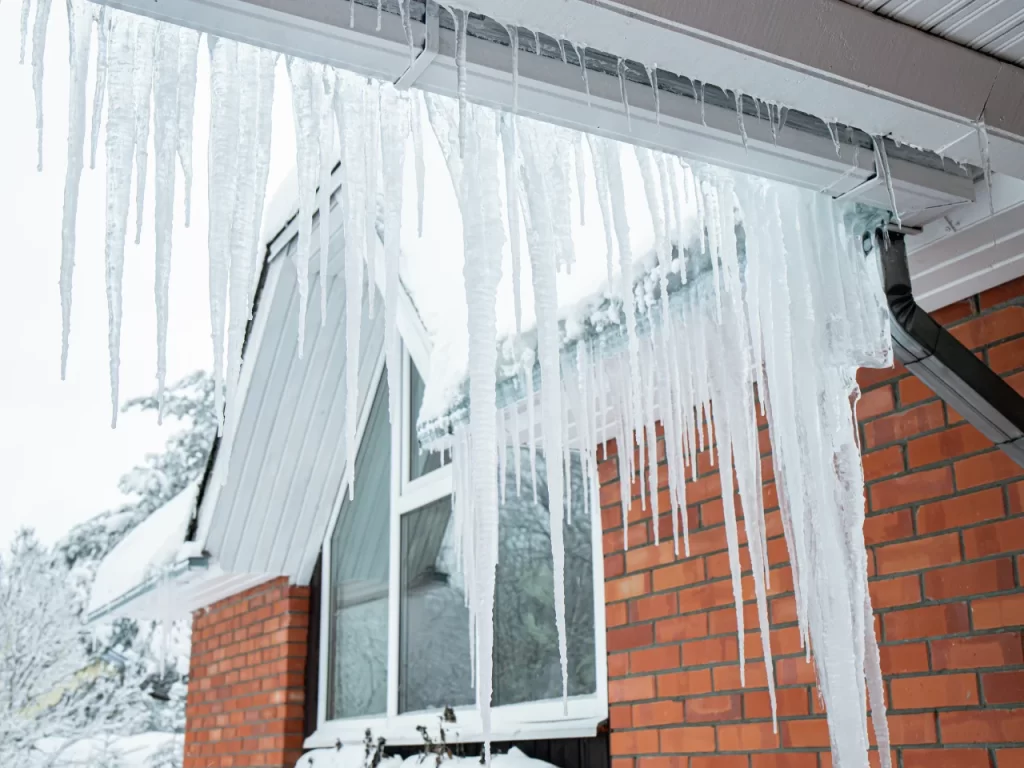 Long, sharp icicles hang from the edge of a roof above a brick house, with snow visible on the roof and in the background, indicating a cold winter day.