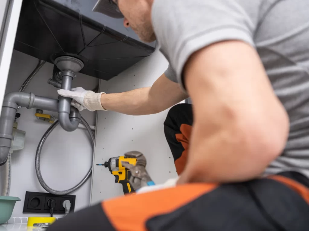 A plumber wearing gloves and work clothes is inspecting or repairing pipes under a sink, with tools and a drill nearby. The scene is inside a cabinet beneath the sink.