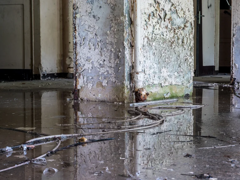 A peeling, weathered column stands in the center of a flooded room, reflecting in the water below. Exposed wires and debris hint at the urgent need for drain repair and plumbing in Toronto on the shiny, damaged floor.