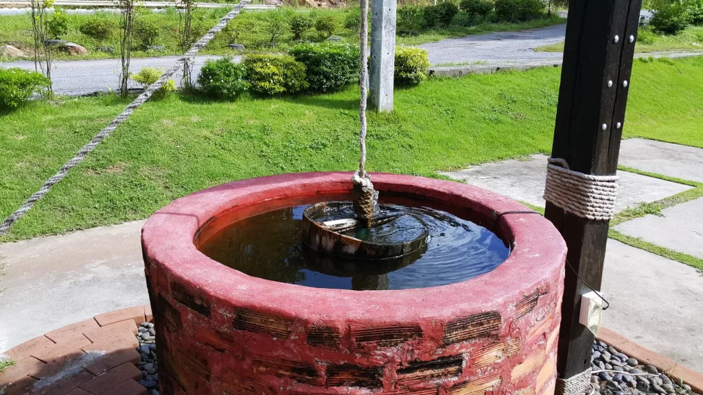 A brick water well with a bucket attached to a rope sits on a lawn near a paved path, with green grass and bushes in the background.