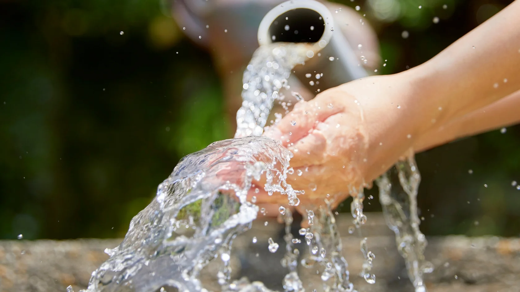 A close-up of hands being washed under a stream of clear water flowing from a pipe, with water droplets splashing and a blurred green background.