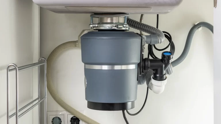 A close-up view of a garbage disposal unit installed under a kitchen sink, showing connected pipes, hoses, and electrical outlets nearby.