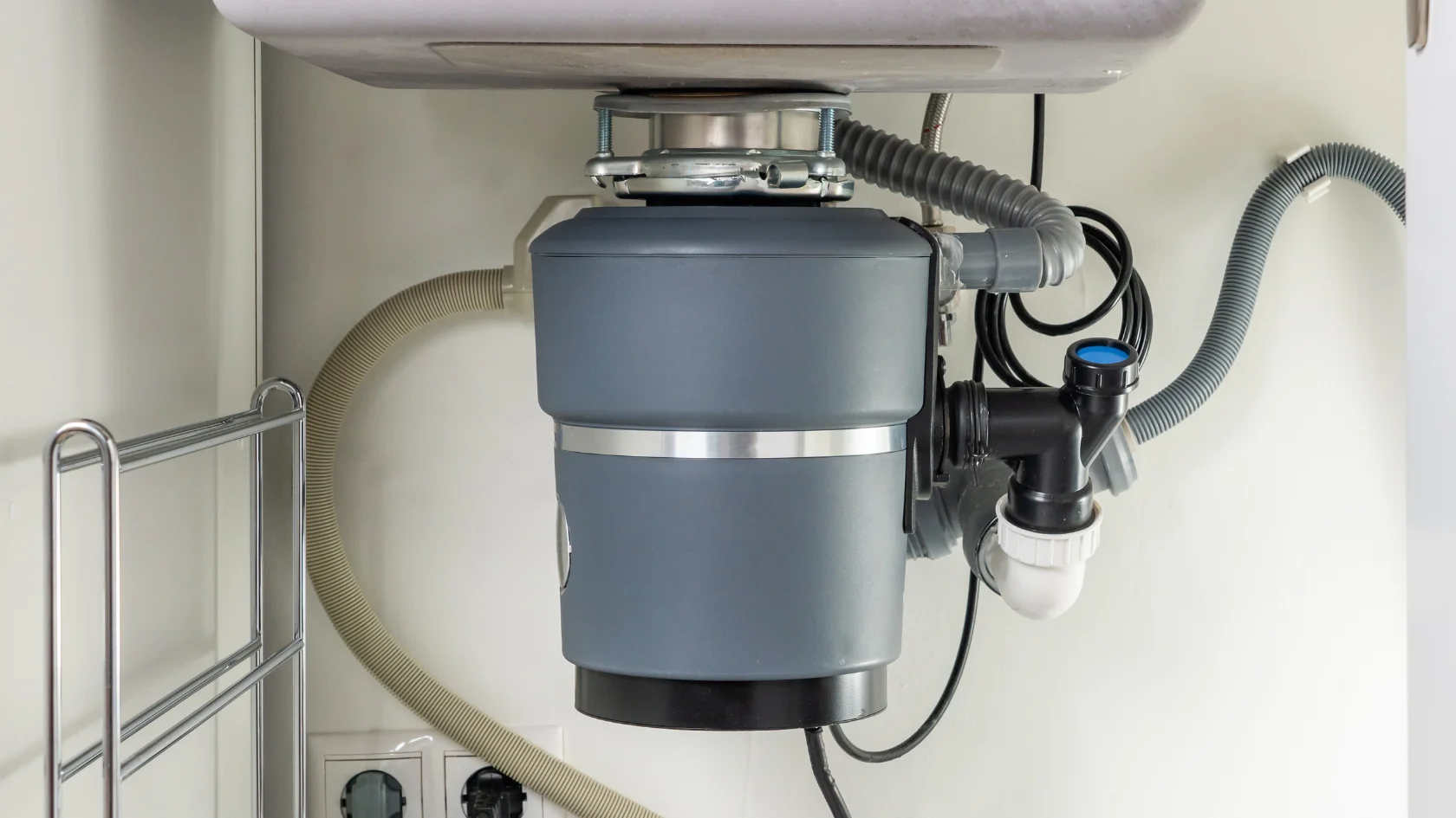 A close-up view of a garbage disposal unit installed under a kitchen sink, showing connected pipes, hoses, and electrical outlets nearby.