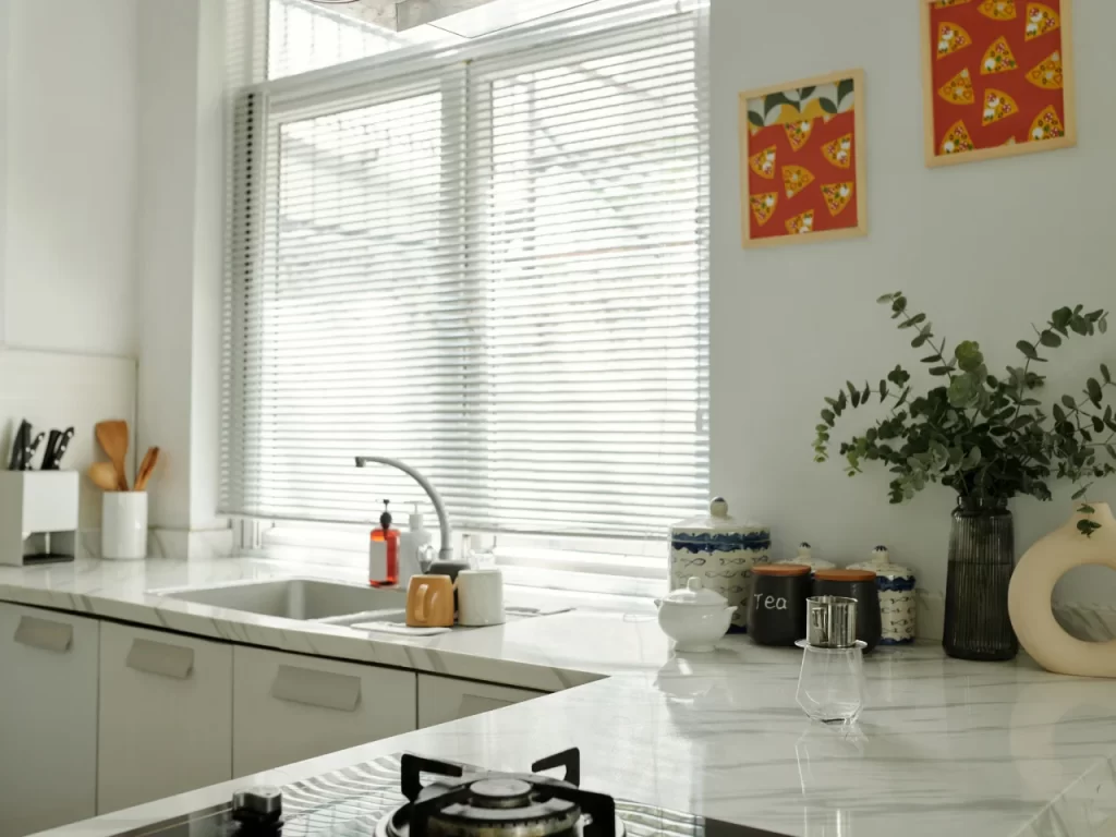 A bright kitchen with white cabinets, marble countertops, a sink, and a stove. There are kitchen items like mugs, jars, and a teapot on the counter, with a plant and colorful wall art above. Blinds cover large windows.