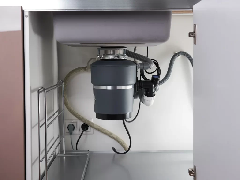 A garbage disposal unit installed under a kitchen sink, with pipes, hoses, and an electrical outlet visible inside the open cabinet.