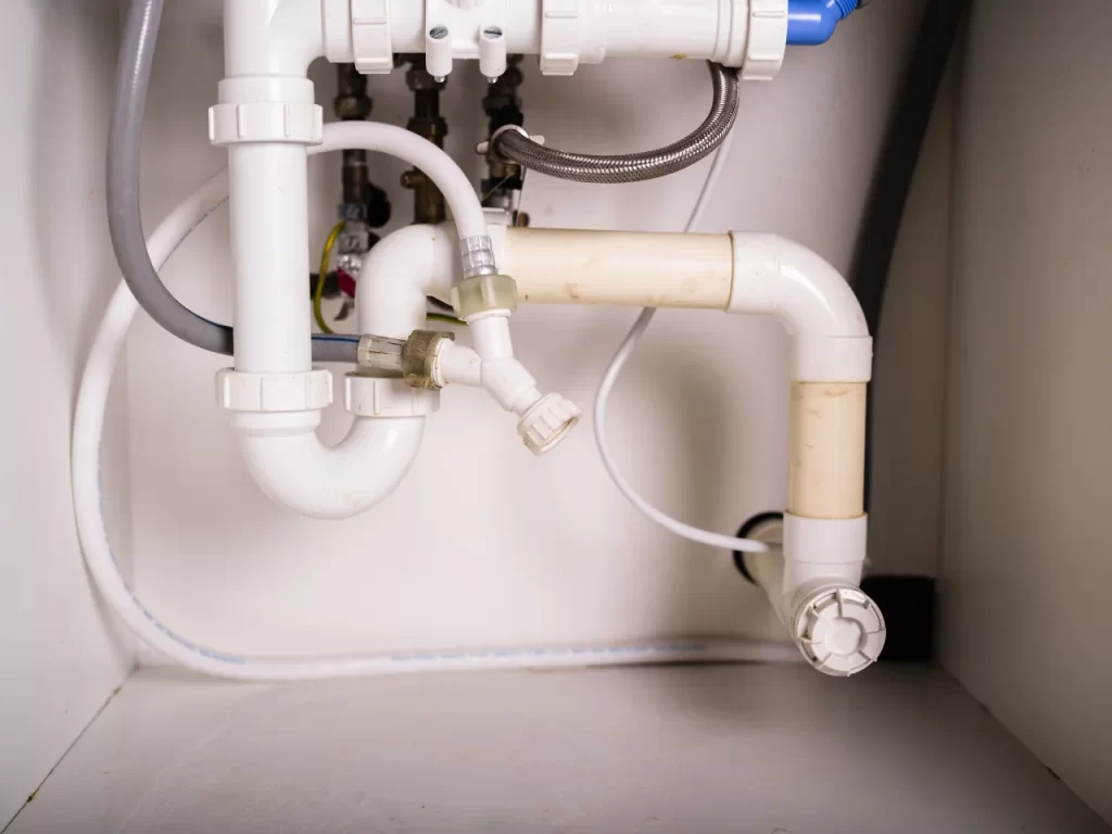 White PVC plumbing pipes and fittings under a sink, including a P-trap and connecting hoses, are shown against a white wall inside a cabinet.