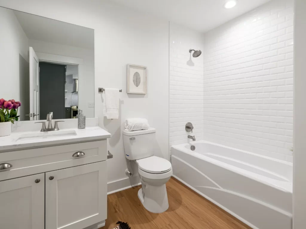 Modern white bathroom with wooden floor, featuring a sink with cabinet, mirror, toilet, towel rack, wall art, and a bathtub with shower. Bright lighting and minimal decor create a clean, fresh look.