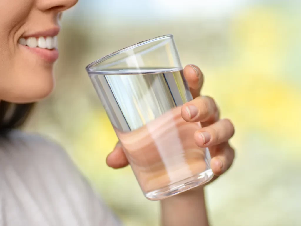 A person smiling and holding a clear glass of water close to their mouth, preparing to take a drink, with a soft, blurred background.