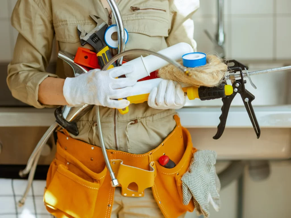 A person wearing a tool belt and gloves holds various plumbing tools and materials, including a caulking gun, hose, tape, and rope, in a kitchen setting.