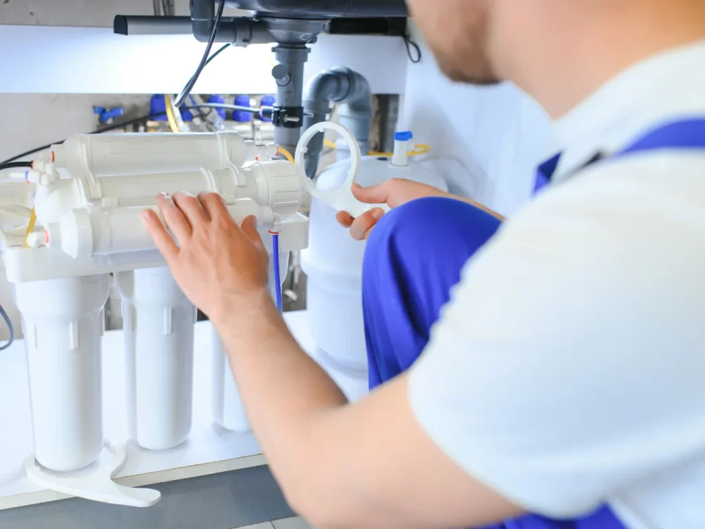 A person wearing a white shirt and blue overalls is kneeling under a sink, inspecting and adjusting a water filtration system with multiple filter cartridges.