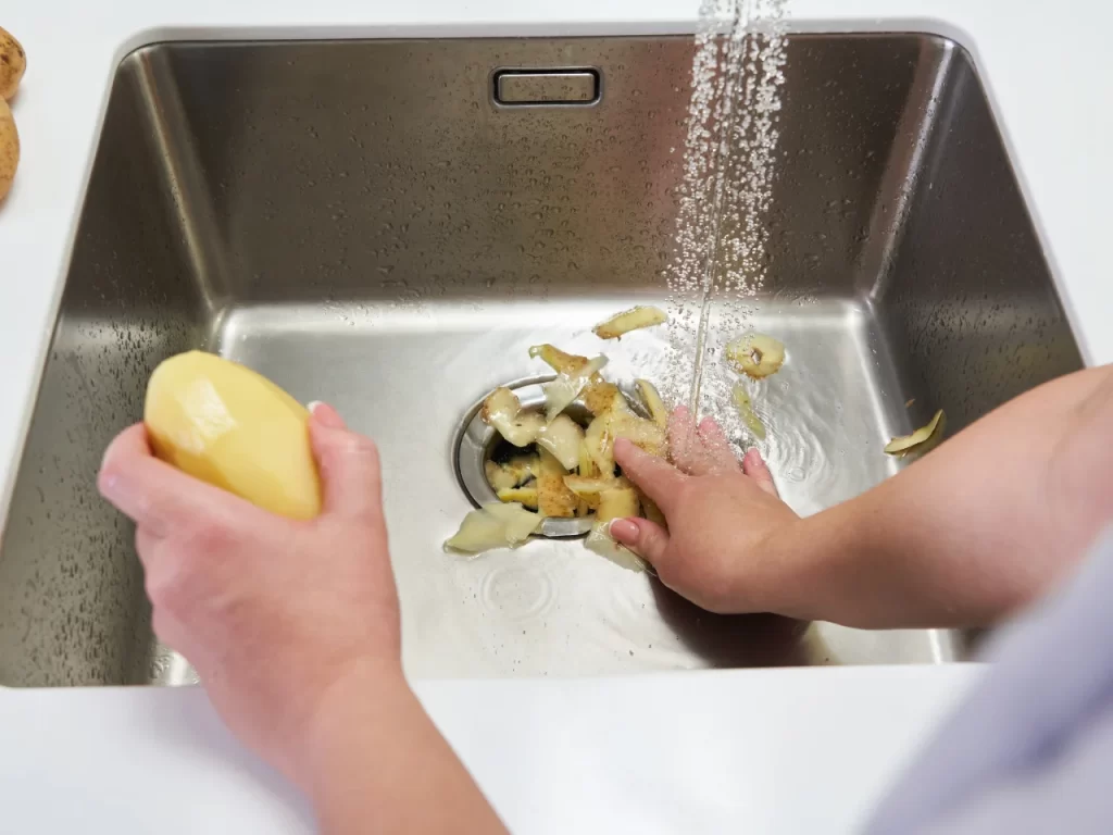 A person holds a peeled potato in one hand and rinses potato peels down a kitchen sink drain with running water using the other hand.