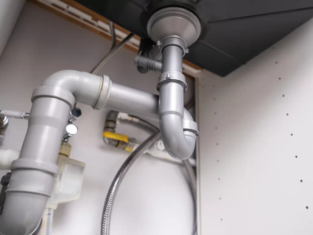 Under-sink view showing a modern plumbing system with grey PVC pipes, a P-trap, and various metal connectors and valves, all installed in a white cabinet.