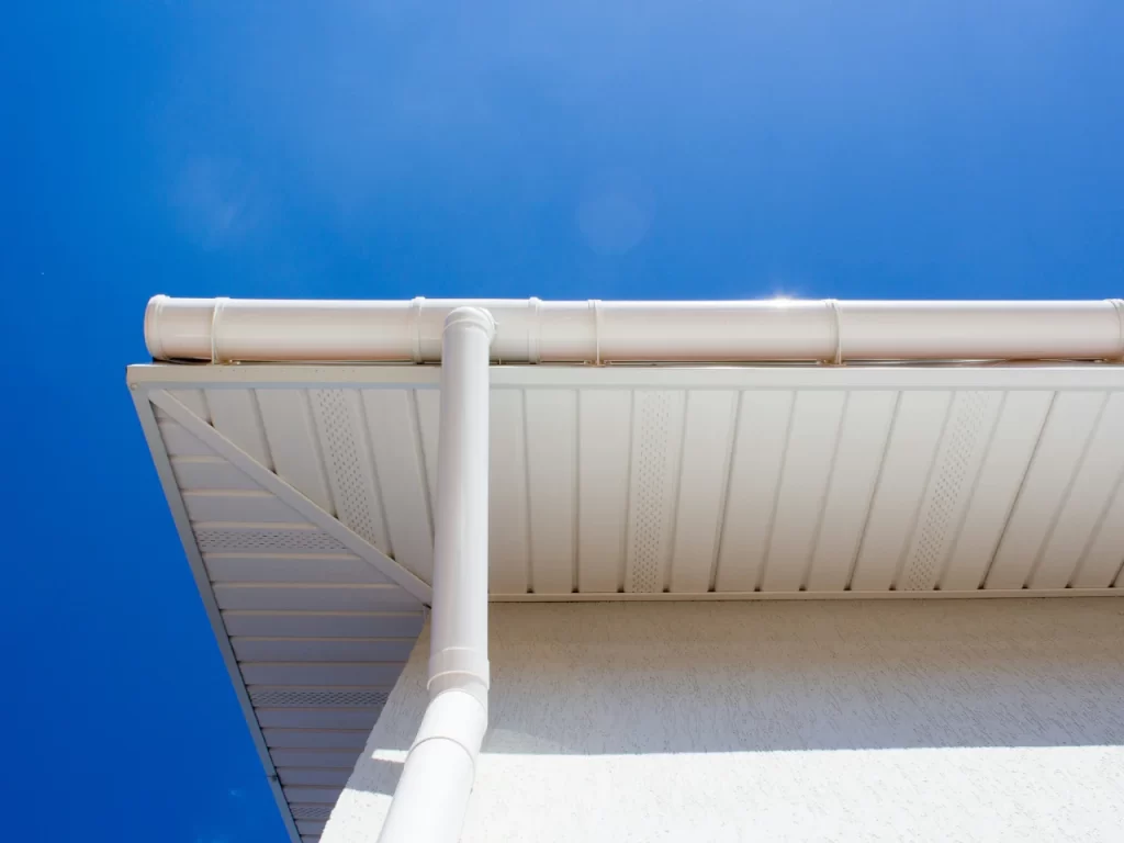 Close-up view of the corner of a house roof with a white rain gutter and downspout, set against a clear blue sky. The buildings soffit and fascia are also visible.