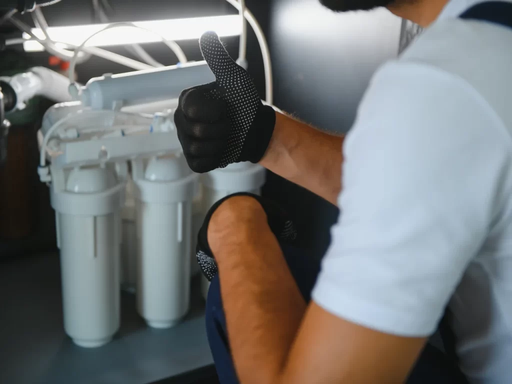 A person wearing black gloves and a white shirt gives a thumbs-up while inspecting or installing a water filtration system under a sink.