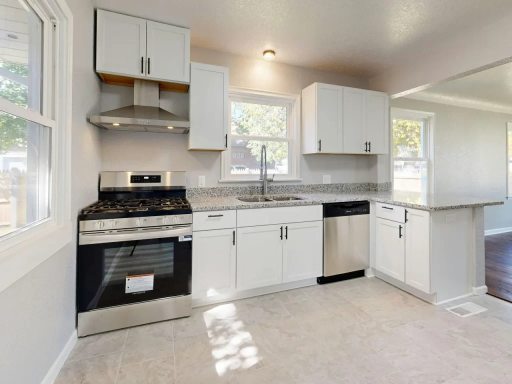 A modern kitchen with white cabinets, a stainless steel gas stove and range hood, granite countertops, a dishwasher, and large windows letting in natural light. The floor is tiled in a light color.