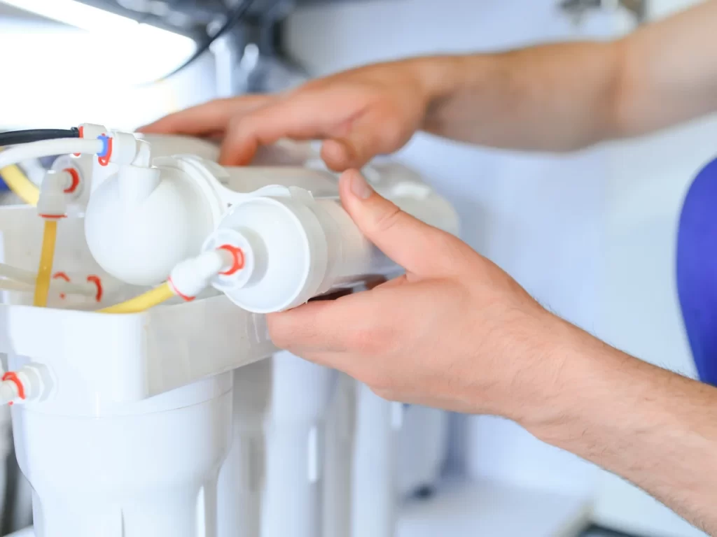 A person’s hands are installing or adjusting a white water filtration system with multiple cylindrical filters and attached tubes. The background is out of focus, highlighting the filtration unit.