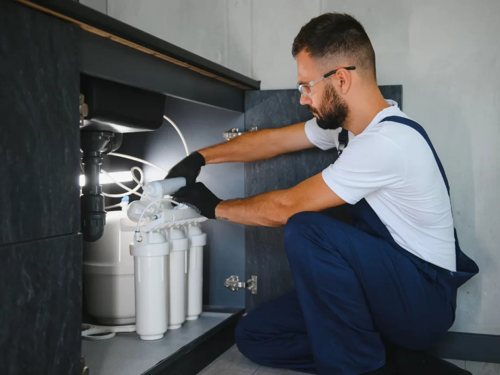 A plumber wearing gloves and protective glasses kneels under a kitchen sink, installing or repairing a water filtration system with multiple white filter cartridges.