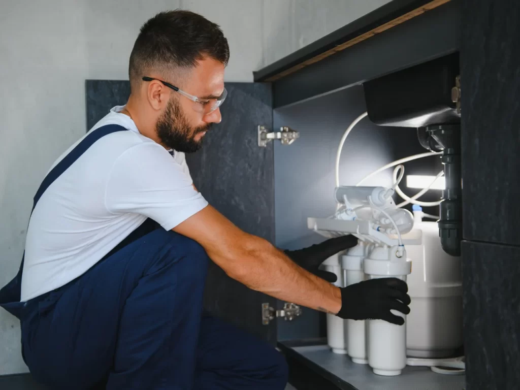 A man wearing glasses, gloves, and overalls installs or repairs a water filtration system under a kitchen sink, with cabinet doors open and pipes visible.