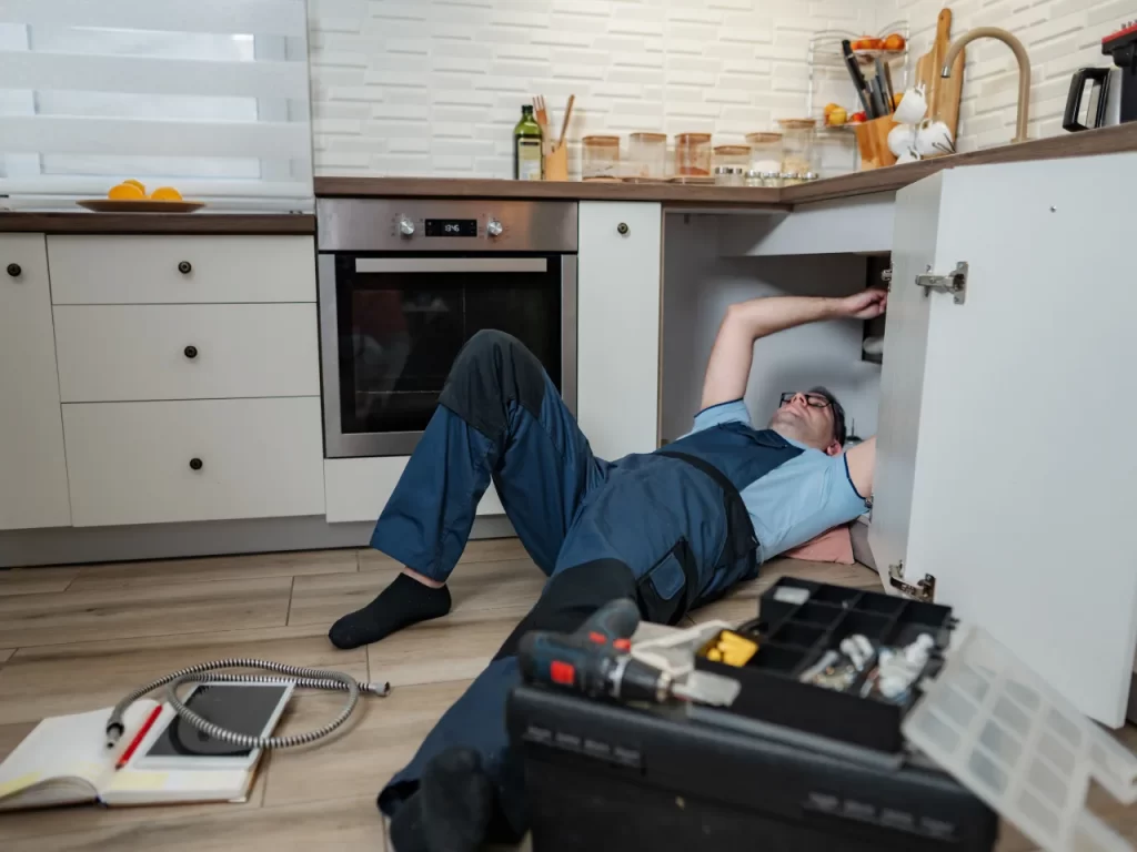 A plumber in overalls lies on the kitchen floor fixing pipes under the sink, surrounded by tools, an open toolbox, and a notebook. The kitchen appears modern and organized.