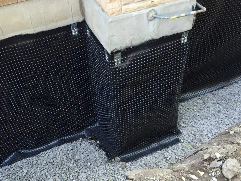 A house foundation wrapped with black dimpled waterproofing membrane, secured with fasteners. Gravel is spread on the ground around the base, part of a concrete block wall is visible above—a common sight during drain repair and plumbing in Toronto.