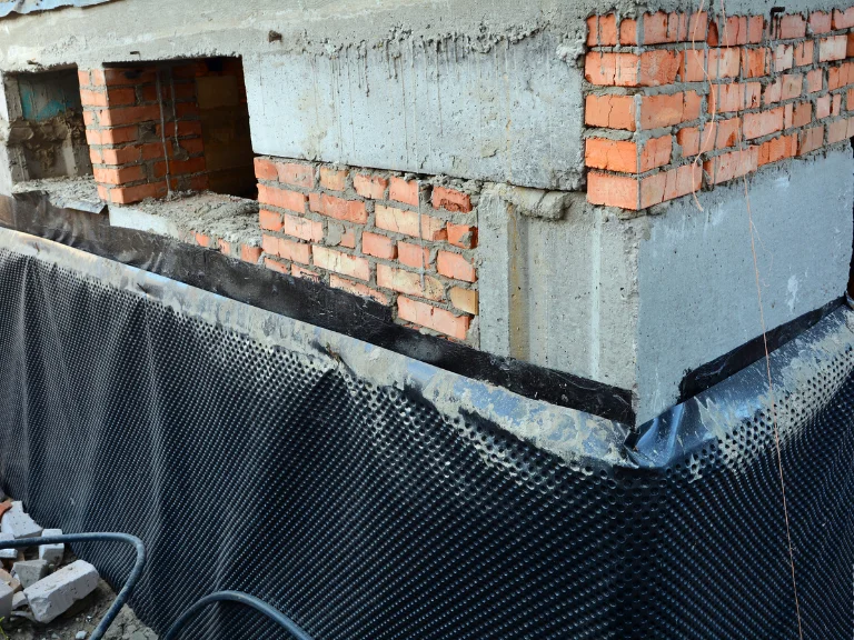 A close-up of a building foundation shows exposed concrete and red brick walls, partially covered with black dimpled waterproofing membrane—highlighting essential work for drain repair and plumbing in Toronto. Construction materials and cables are visible nearby.