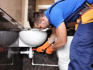 A plumber wearing blue overalls and orange gloves is using a wrench to fix pipes under a white bathroom sink next to a toilet, showcasing expert drain repair and plumbing in Toronto. Tools are visible in the plumber's belt.