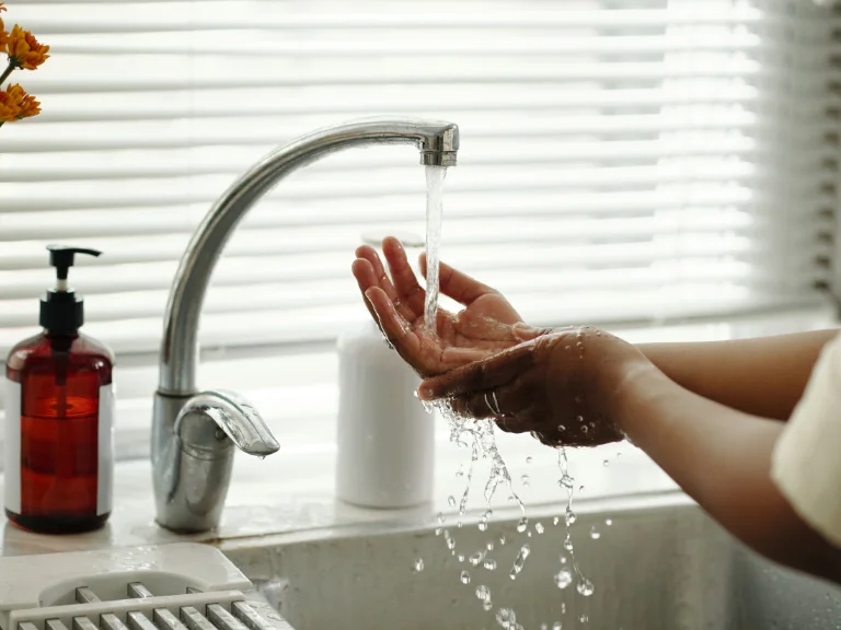 A person washes their hands under a running kitchen faucet, with water splashing—a reminder of the importance of plumbing in Toronto. A red soap dispenser and blinds covering a bright window are in the background.