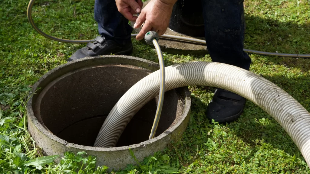 A person holding hoses over an open outdoor septic tank, preparing for cleaning or pumping, standing on green grass.