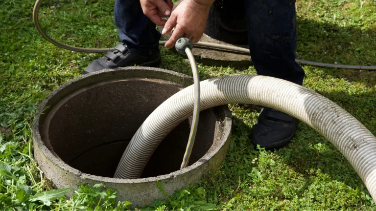 A person holding hoses over an open outdoor septic tank, preparing for cleaning or pumping, standing on green grass.