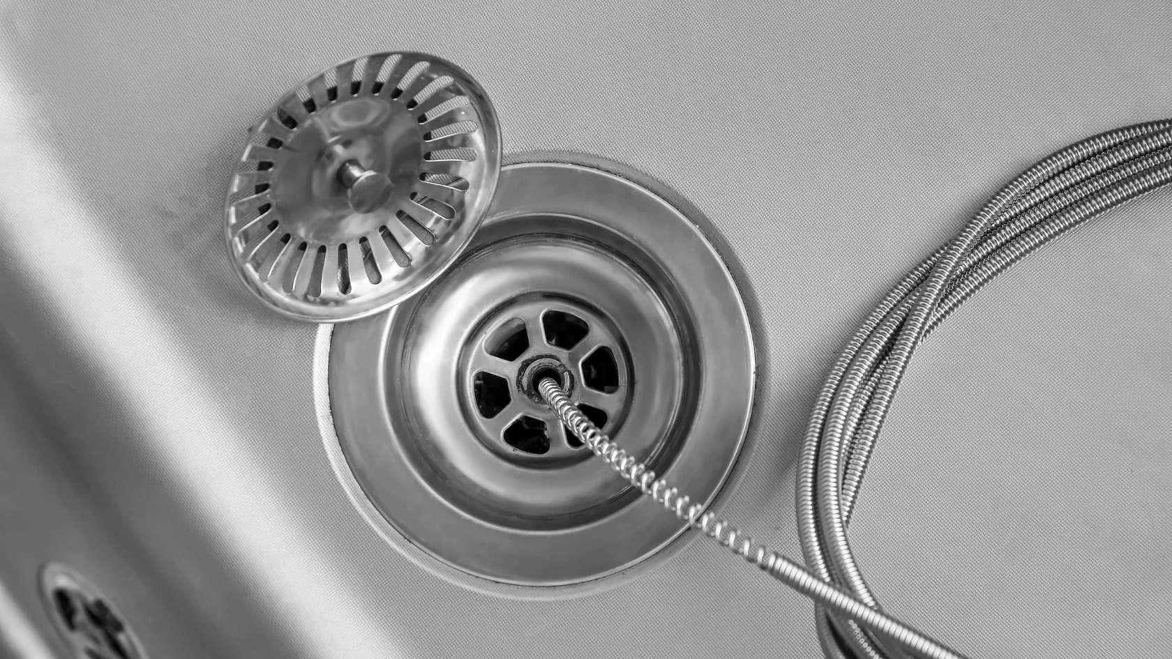 Close-up of a metal kitchen sink drain with the strainer cover removed and a flexible metal drain snake inserted into the drain opening, suggesting that the drain is being unclogged or cleaned.