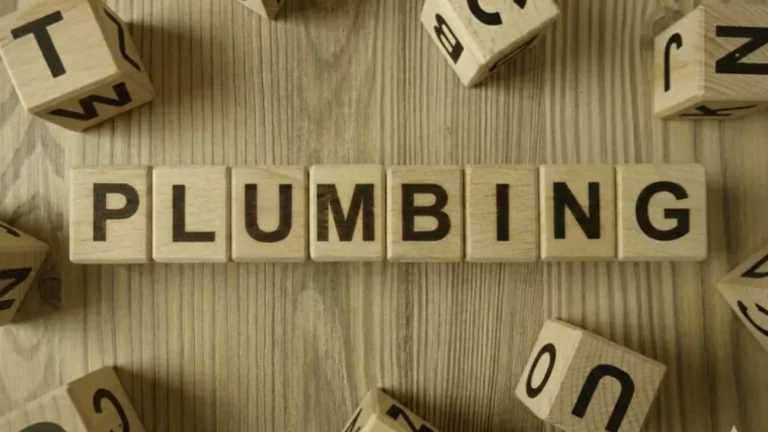 Wooden letter tiles spell PLUMBING on a wooden surface, surrounded by scattered wooden cubes with various letters visible on their sides.
