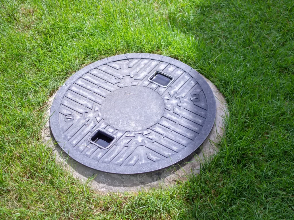 A round, black manhole cover with textured patterns and two rectangular openings, set in a concrete rim and surrounded by green grass.