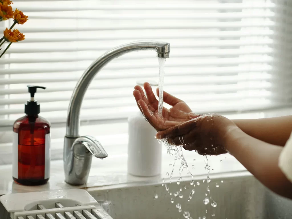 A person washes their hands under running water from a kitchen faucet, with splashing water droplets visible. A soap dispenser, flowers, and a window with blinds are in the background.