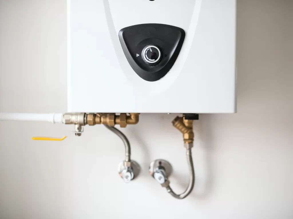 Close-up of a white wall-mounted water heater with visible pipes, valves, and connectors below it, attached to a white wall. The unit features a black control knob in the center.