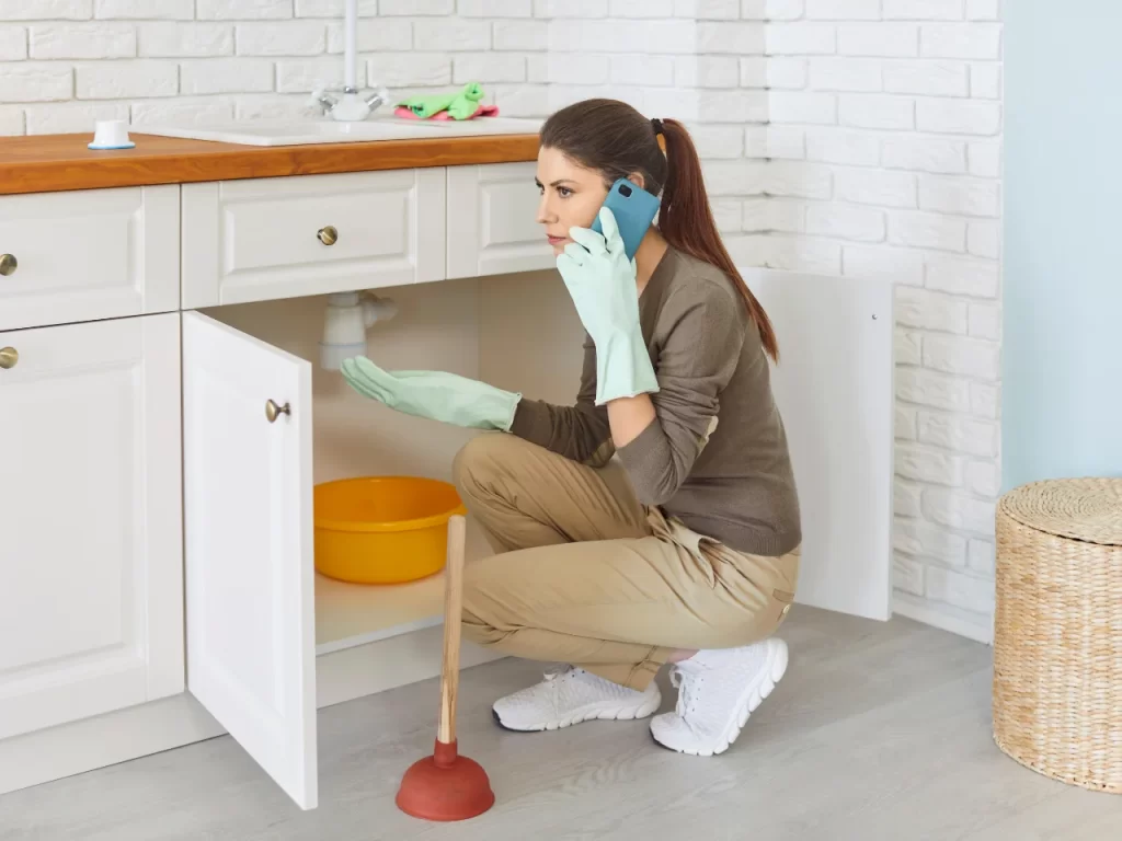 A woman wearing gloves and holding a plunger talks on the phone while inspecting a leak under a kitchen sink, with a bucket placed below the pipes.