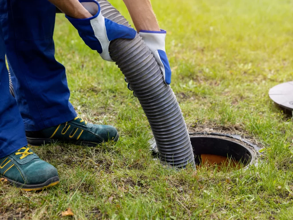 A person wearing gloves and work clothes holds a large hose, placing it into an open outdoor septic tank or manhole on a grassy lawn for maintenance or cleaning.