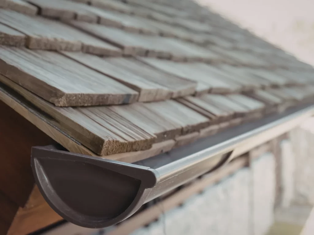 Close-up view of a house roof with wooden shingles and a metal rain gutter attached along the edge for water drainage. The image focuses on the shingles and gutter in natural light.
