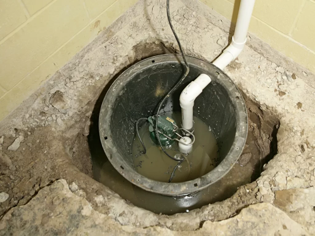 A sump pump installed in a basement floor pit with standing water, surrounded by cracked and damaged concrete and connected to white PVC pipes.