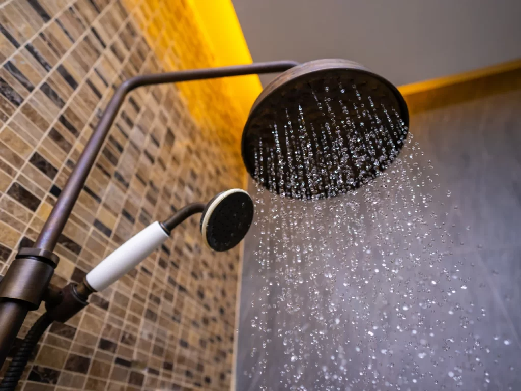 A close-up of a modern showerhead releasing water, with a handheld shower attachment nearby. The background features brown mosaic tiles and warm yellow lighting at the ceiling.