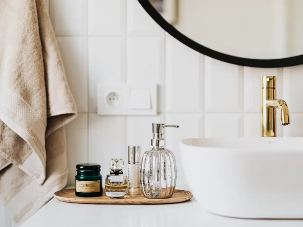 A bathroom counter with a white sink, gold faucet, soap dispenser, skincare jars, and perfume bottles on a wooden tray. A beige towel hangs on the wall beside white tiles and a round mirror.