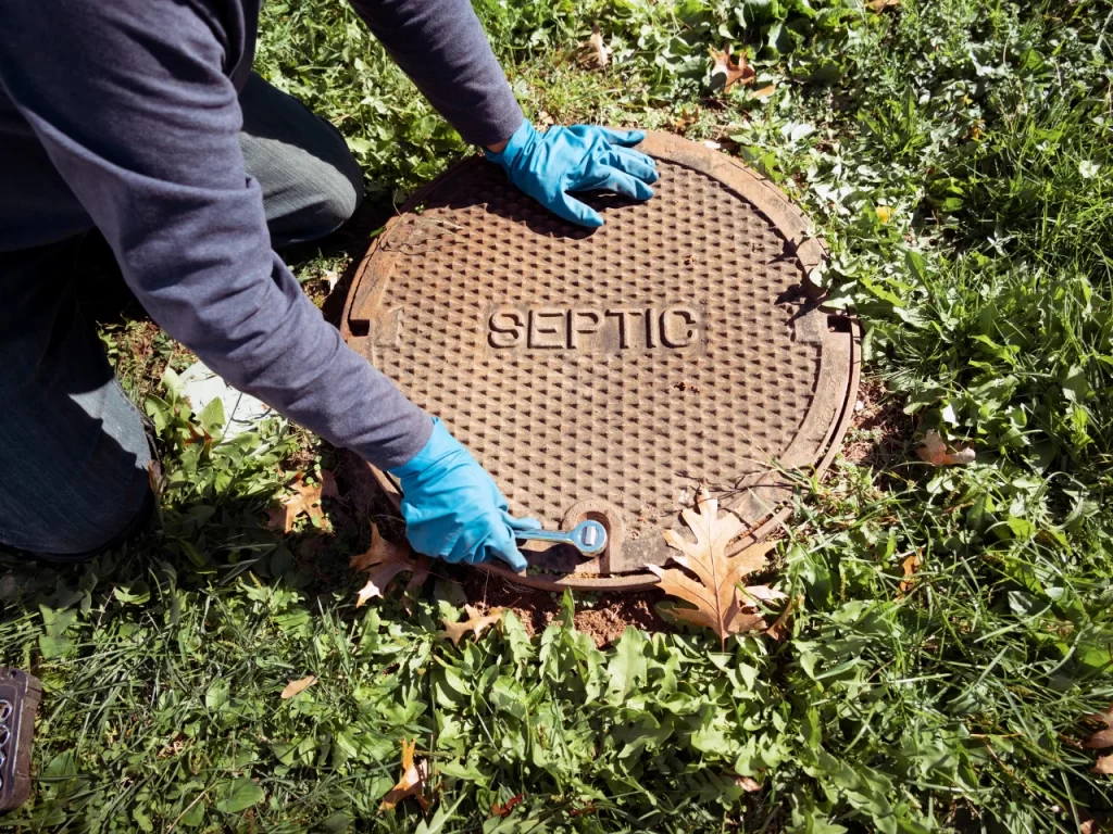 A person wearing blue gloves and a dark sweatshirt uses a tool to lift a round, metal septic tank cover, surrounded by green grass and fallen leaves.