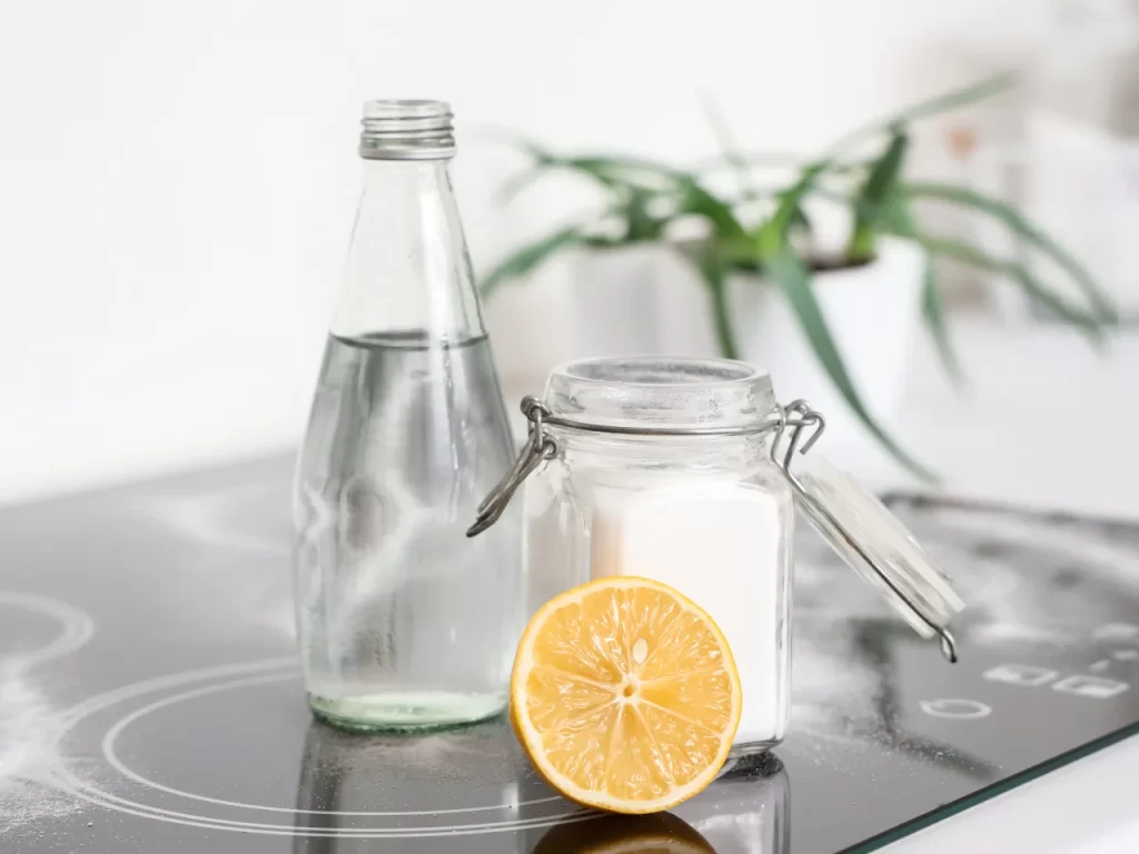 A glass bottle of clear liquid, a glass jar of white powder, and a halved lemon are arranged on a kitchen counter with a potted plant in the background.