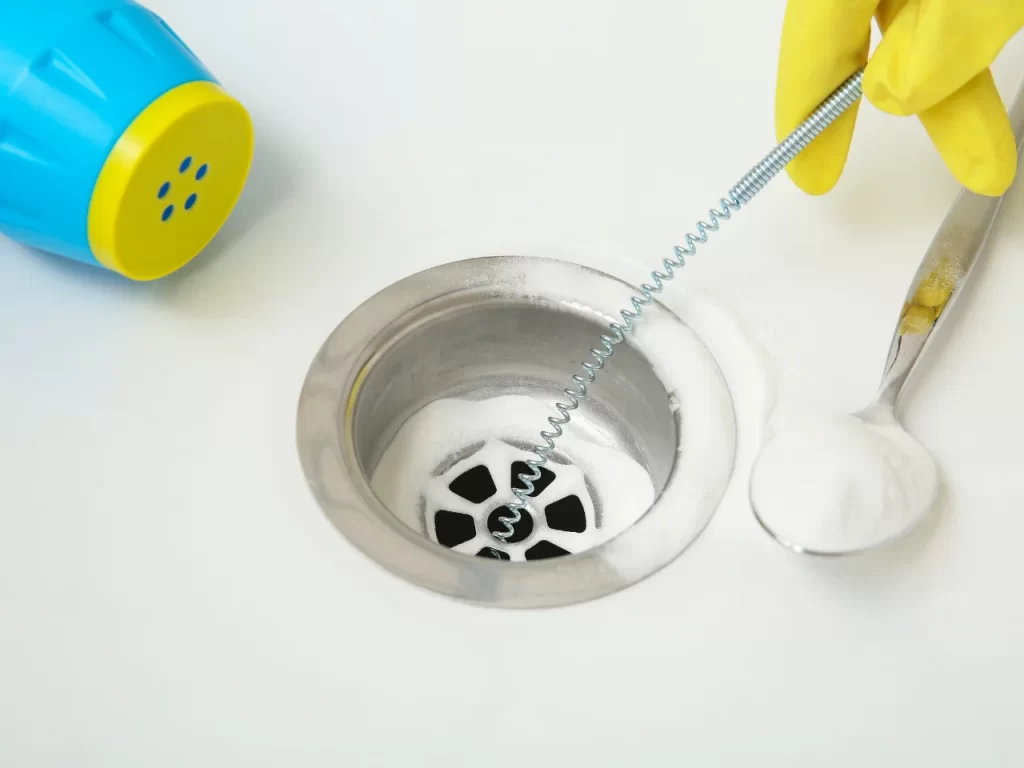 A person wearing yellow gloves uses a drain snake tool to clean a kitchen sink drain, with baking soda and a spoon nearby on a white countertop.