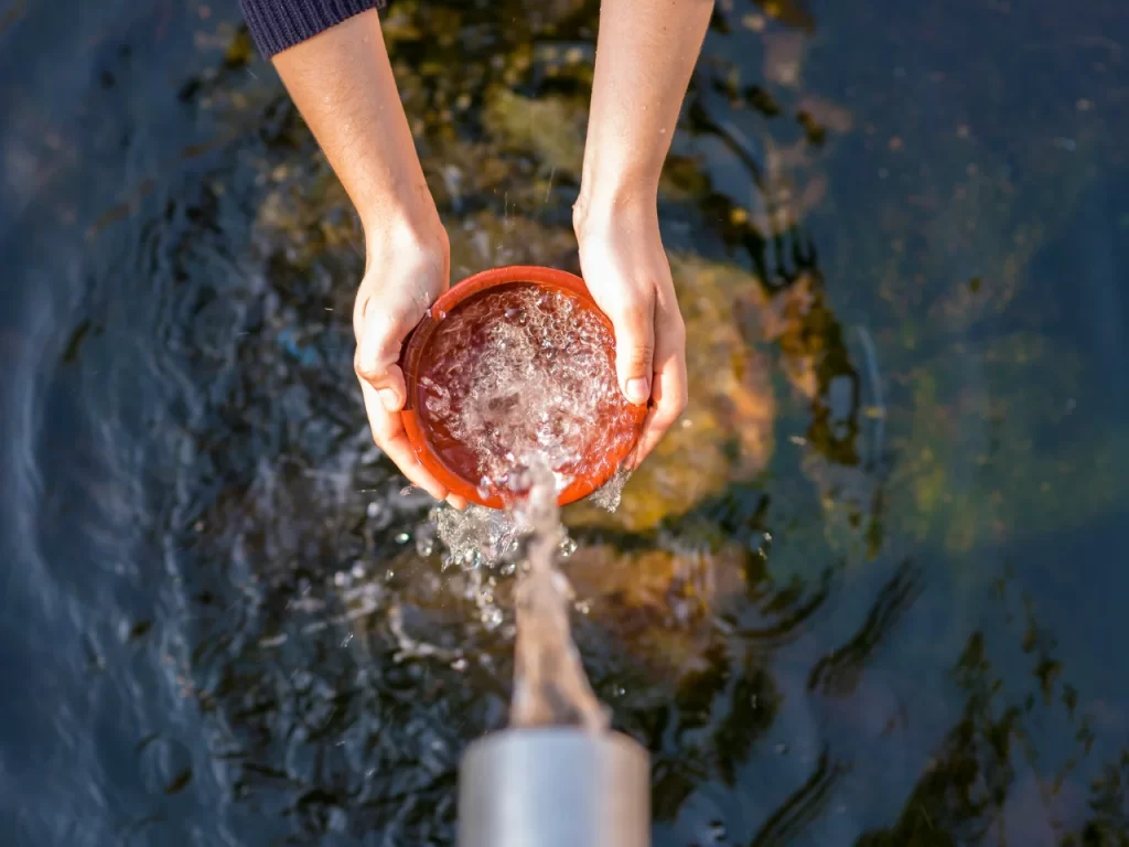 A person holds a red bowl under a stream of water outdoors, filling it while water splashes. The background shows rippling water and the persons hands and arms are visible.