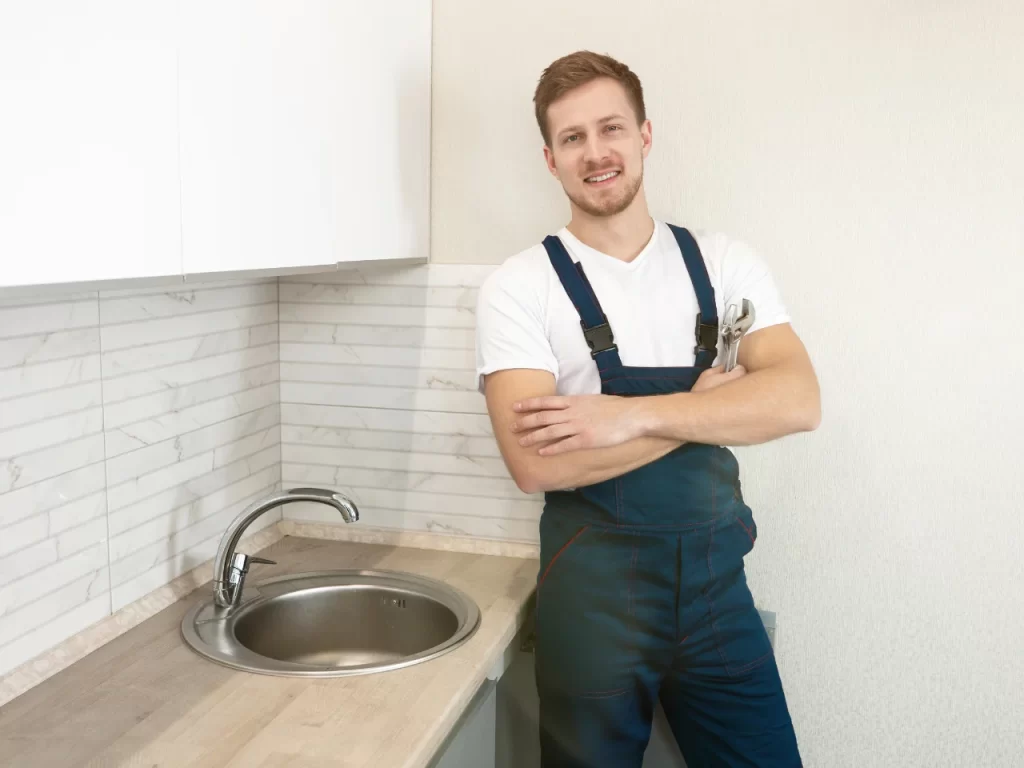 A smiling plumber in blue overalls and a white t-shirt stands with arms crossed next to a kitchen sink, with a wrench tucked into his overalls pocket. The kitchen has light countertops and tiled backsplash.