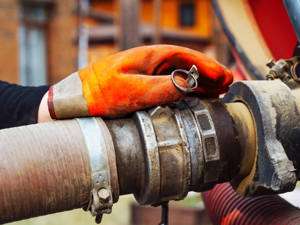 A close-up of a gloved hand with an orange work glove resting on a large industrial hose and metal coupling. The background is slightly out of focus, showing a building structure.
