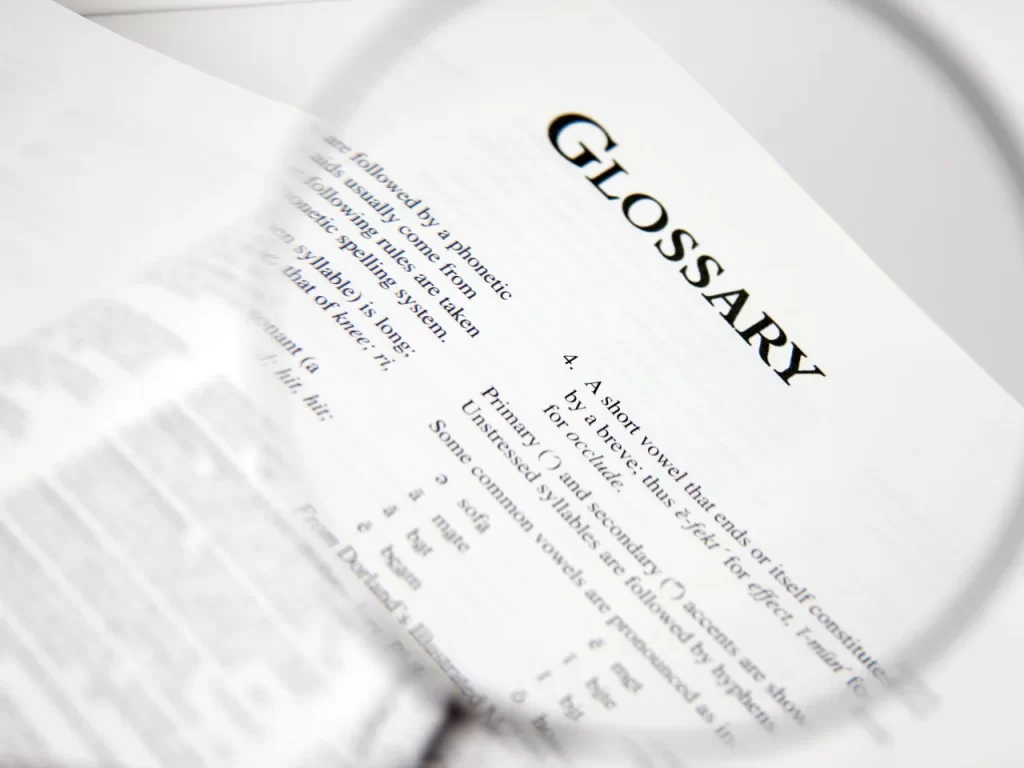 A close-up of a glossary page in a book, with the word GLOSSARY in bold at the top. A magnifying glass is partially visible, enlarging some of the printed text below the heading.