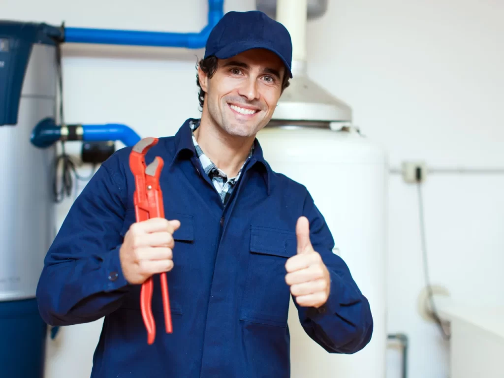 A smiling plumber in a blue uniform and cap holds a red pipe wrench in one hand and gives a thumbs-up with the other, standing in front of plumbing equipment indoors.