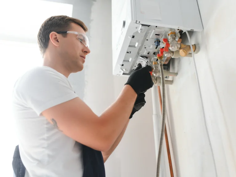 A person wearing safety glasses and gloves is servicing or repairing a wall-mounted boiler or heating system, using a tool to work on pipes and valves—ideal for drain repair and plumbing in Toronto.