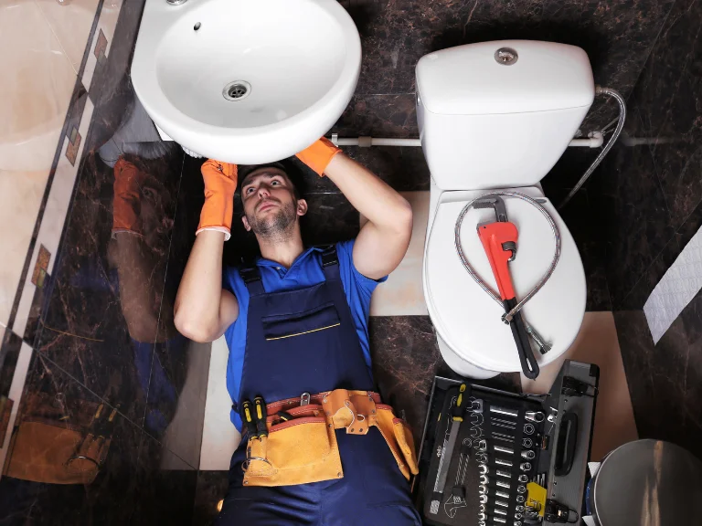 A plumber in blue overalls and orange gloves lies under a bathroom sink, fixing plumbing. Tools, including a large wrench and toolbox, are near the toilet—perfect for expert drain repair and plumbing in Toronto.